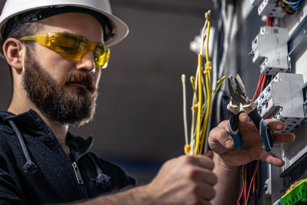 A male electrician works in a switchboard with an electrical connecting cable, connects the equipment with tools.